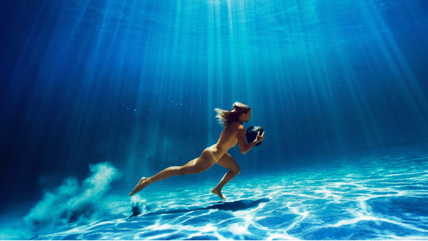a female athlete walks on the ocean floor carrying a large rock with sunlight shining down from above the water
